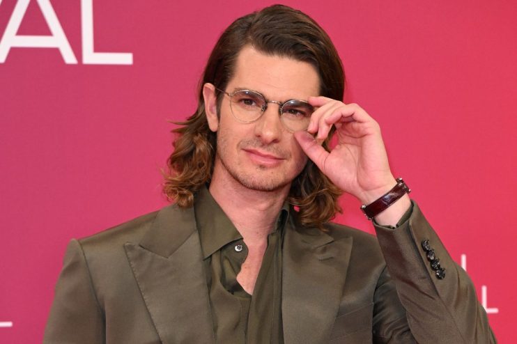 Andrew Garfield in a green suit and glasses, hand adjusting his spectacles, against a pink background for AI film news.