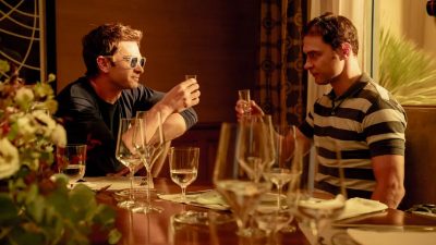 Andrew Garfield (left) in sunglasses and another man toast with shot glasses at a dinner table.