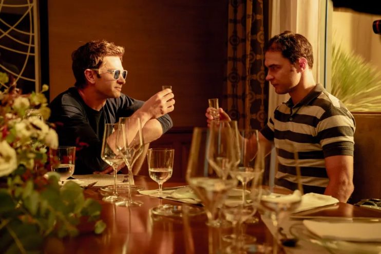 Andrew Garfield (left) in sunglasses and another man toast with shot glasses at a dinner table.