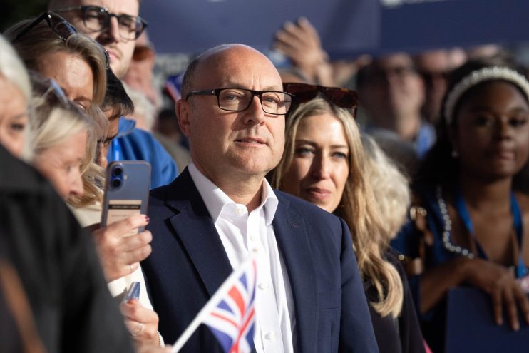 Andrew Griffith, Conservative MP, in glasses and dark suit, holding a Union Jack flag at an event.