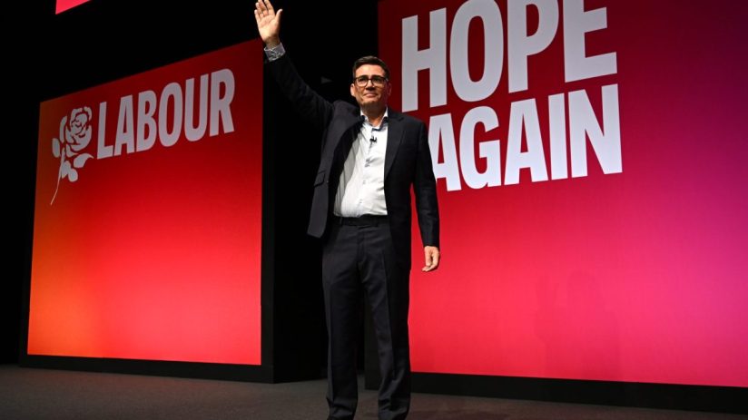 Andy Burnham waving on stage at a Labour Party Conference with HOPE AGAIN slogan visible