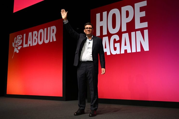 Andy Burnham waving on stage at a Labour Party Conference with HOPE AGAIN slogan visible