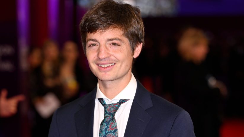 Simon Rich smiling in a dark suit and patterned tie, with a blurred purple background