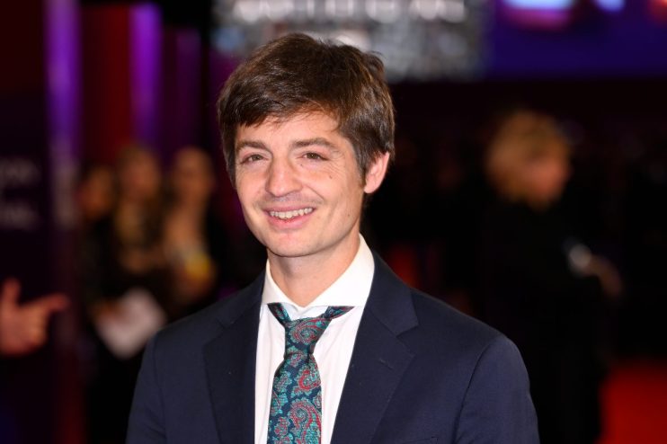 Simon Rich smiling in a dark suit and patterned tie, with a blurred purple background