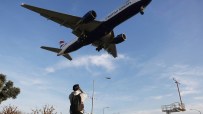 British Airways jet, landing gear down, flying low over a man taking a photo, with clear blue sky background.