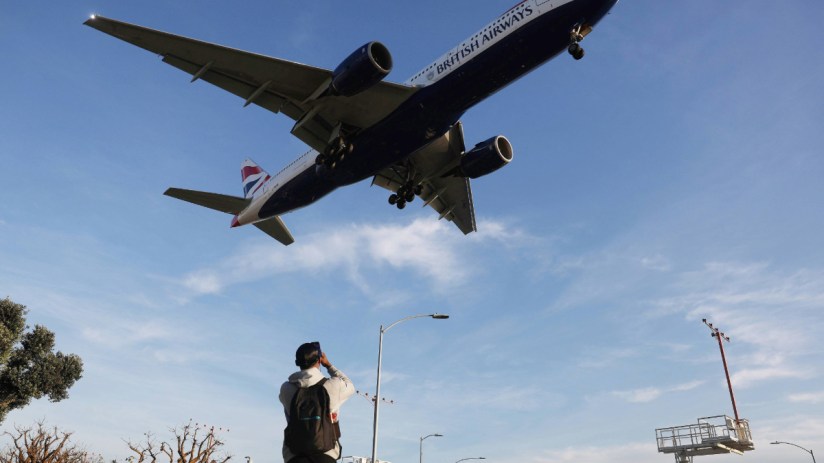 British Airways jet, landing gear down, flying low over a man taking a photo, with clear blue sky background.