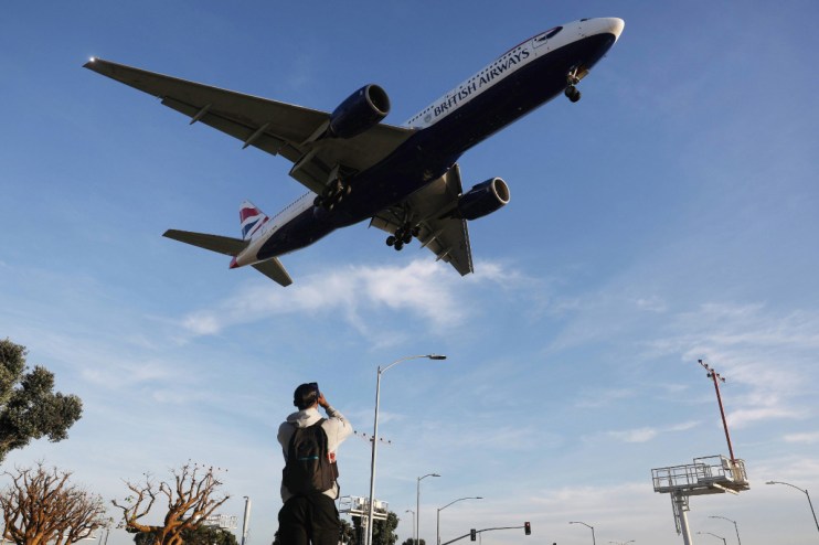British Airways jet, landing gear down, flying low over a man taking a photo, with clear blue sky background.
