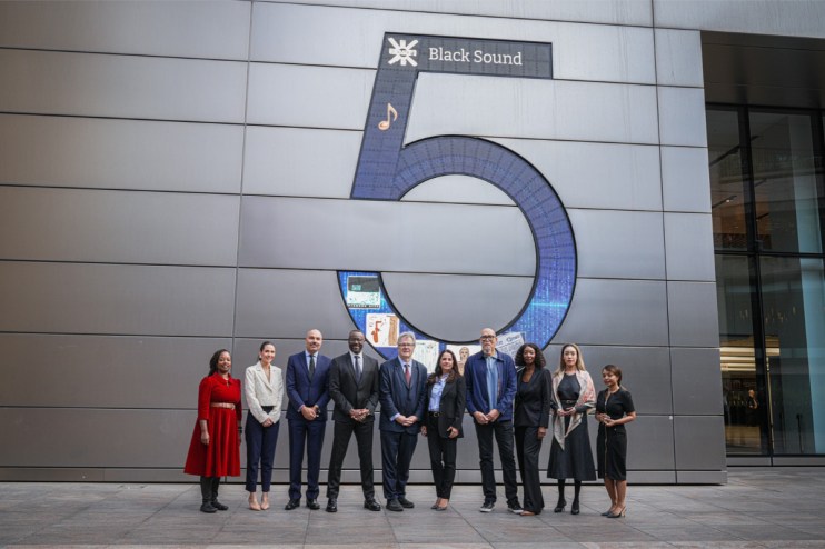 Group of diverse professionals in business attire standing in front of a UBS building with Black Sound 5 display for Black...