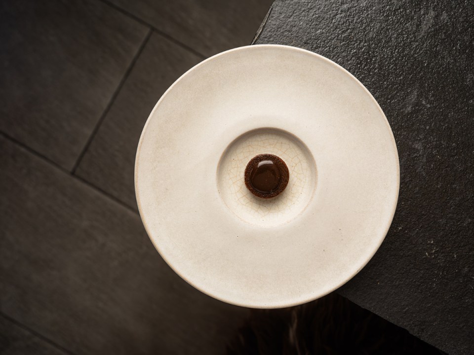 A plain white plate with a small round chocolate dessert in the center, set against a dark stone surface.