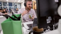 Man with headphones making a face, eating pickles from a jar at his office desk. Budget 2026.