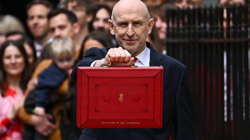 Healey holding the red Budget Box, smiling at the camera with a crowd visible in the background.