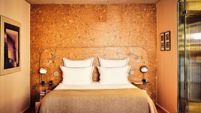 Hotel room with cork wall, bed with white pillows and brown blanket, modern lamps, and framed art.