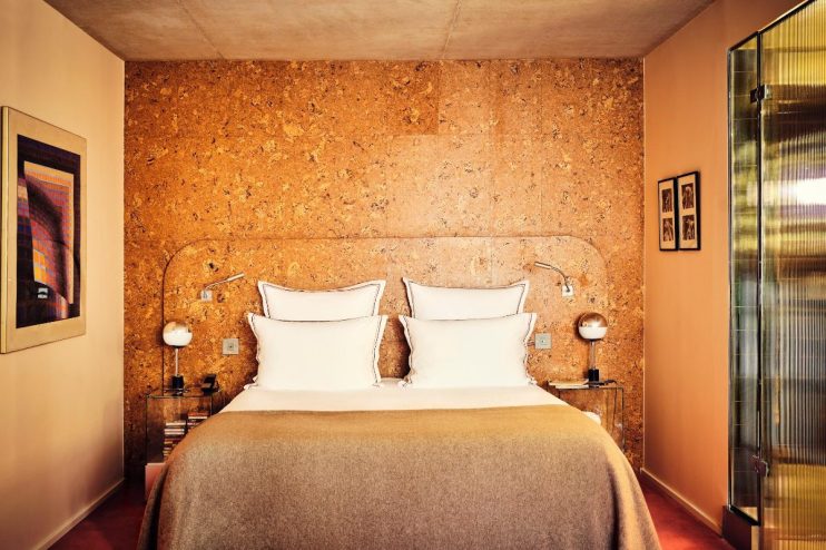 Hotel room with cork wall, bed with white pillows and brown blanket, modern lamps, and framed art.