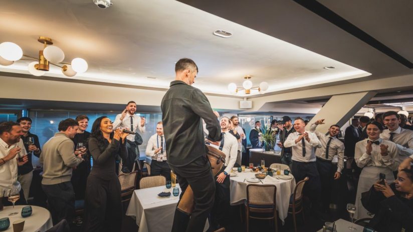 Guests and staff cheering, clapping, and singing at a lively event in a restaurant dining room.