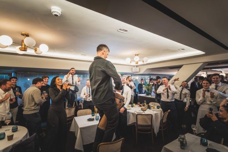 Guests and staff cheering, clapping, and singing at a lively event in a restaurant dining room.