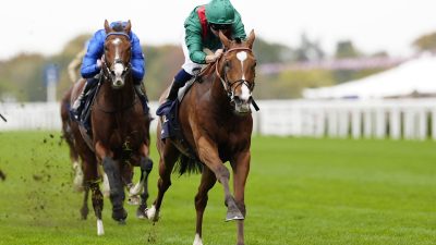 Two racehorses and jockeys in green and blue silks competing on a green turf track.