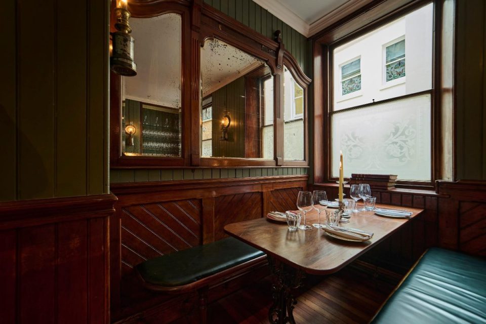 Cozy restaurant interior with dark wood paneling, green walls, antique mirrors, and a set table by a window.