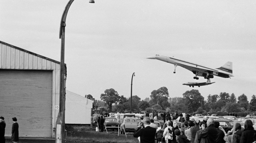 Black and white Concorde supersonic jet flying low over a crowd of onlookers and parked cars in 1969.