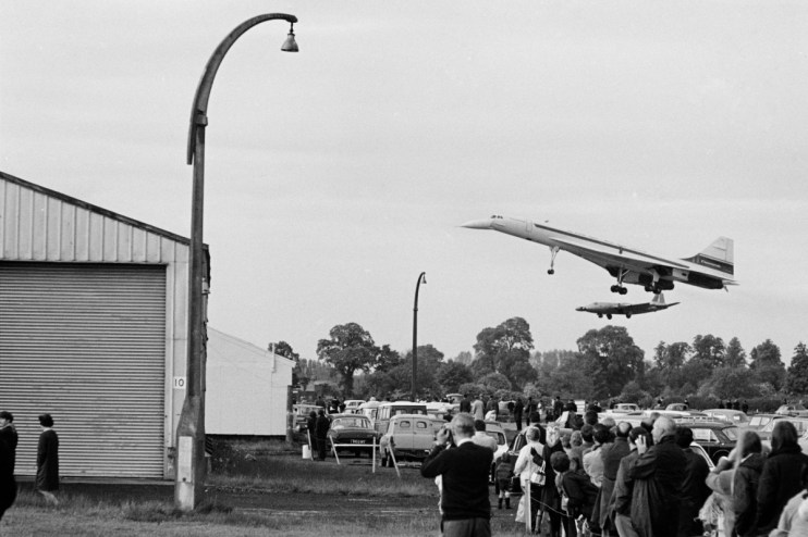 Black and white Concorde supersonic jet flying low over a crowd of onlookers and parked cars in 1969.