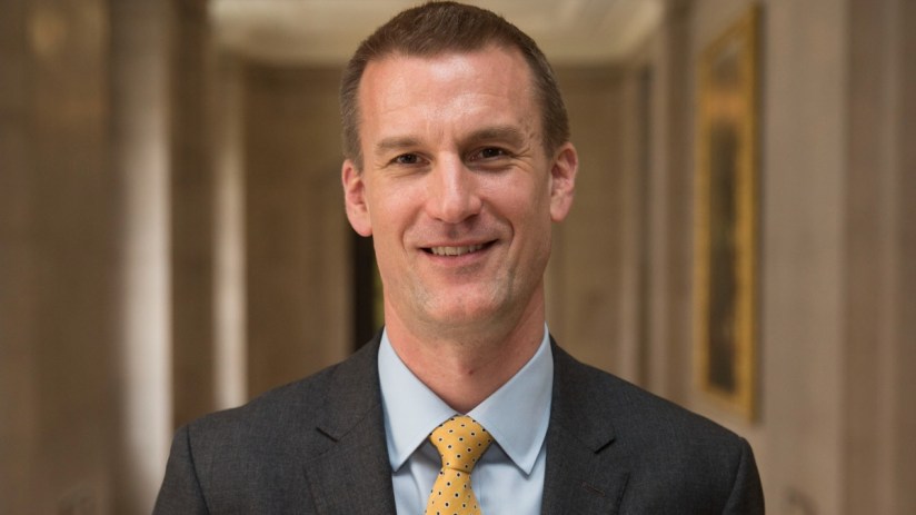 David Bailery, a man in a suit and yellow tie, smiling in a well-lit hall with a framed picture in the background.