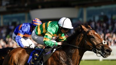 Jockey in green and yellow silks on a brown racehorse, with another jockey in blue behind, on a sunny track