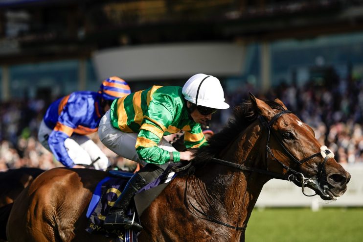 Jockey in green and yellow silks on a brown racehorse, with another jockey in blue behind, on a sunny track