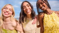 Three smiling women in sequined dresses at a beach, under a blue sky, representing Debenhams fashion.