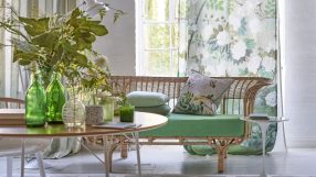 Designers Guild interior with rattan sofa, green cushions, floral and striped curtains, and a wooden table with foliage.