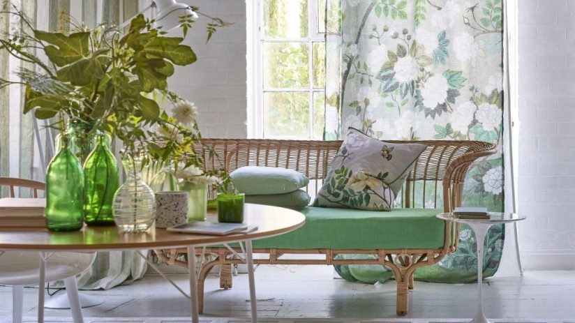 Designers Guild interior with rattan sofa, green cushions, floral and striped curtains, and a wooden table with foliage.