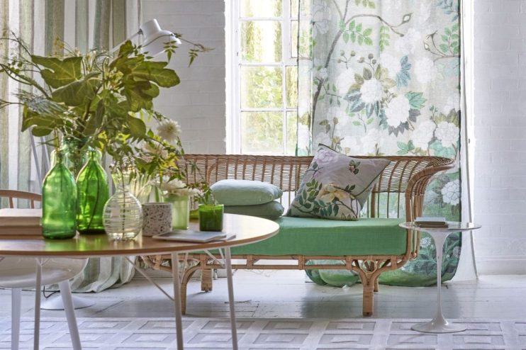 Designers Guild interior with rattan sofa, green cushions, floral and striped curtains, and a wooden table with foliage.