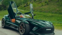 Dark green Donkervoort sports car with orange interior, open doors, parked on a road with a grassy hill background.