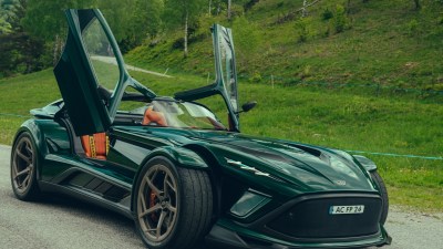 Dark green Donkervoort sports car with orange interior, open doors, parked on a road with a grassy hill background.