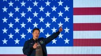 Elon Musk speaking into a microphone, pointing upwards, in front of a large American flag with stars and stripes