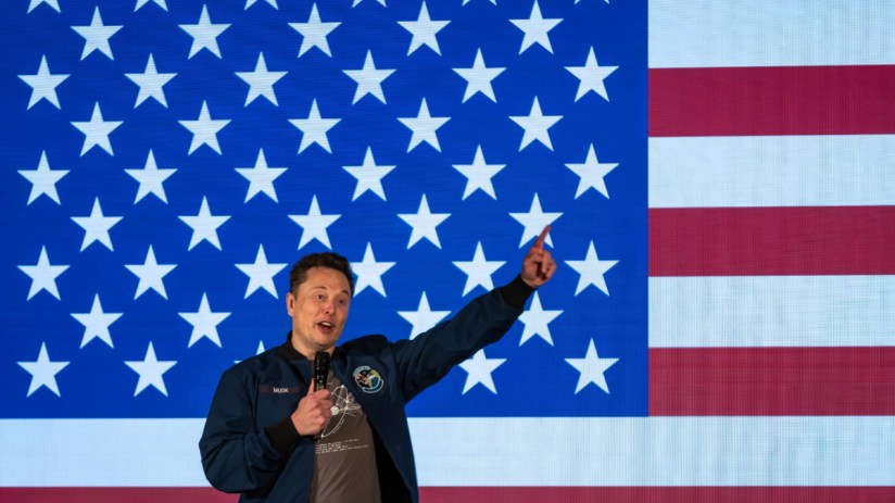 Elon Musk speaking into a microphone, pointing upwards, in front of a large American flag with stars and stripes