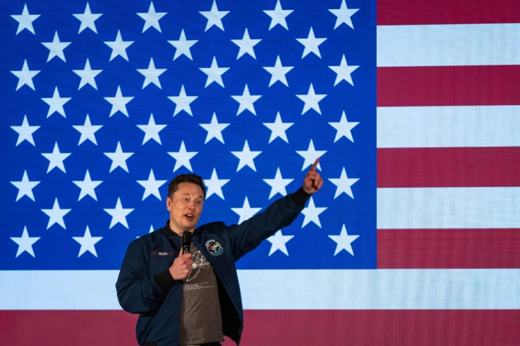 Elon Musk speaking into a microphone, pointing upwards, in front of a large American flag with stars and stripes