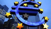 Illuminated Euro symbol with gold stars against a cityscape and blue sky, representing the Eurozone
