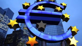 Illuminated Euro symbol with gold stars against a cityscape and blue sky, representing the Eurozone