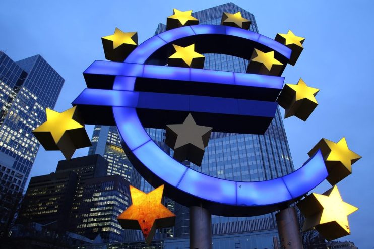 Illuminated Euro symbol with gold stars against a cityscape and blue sky, representing the Eurozone