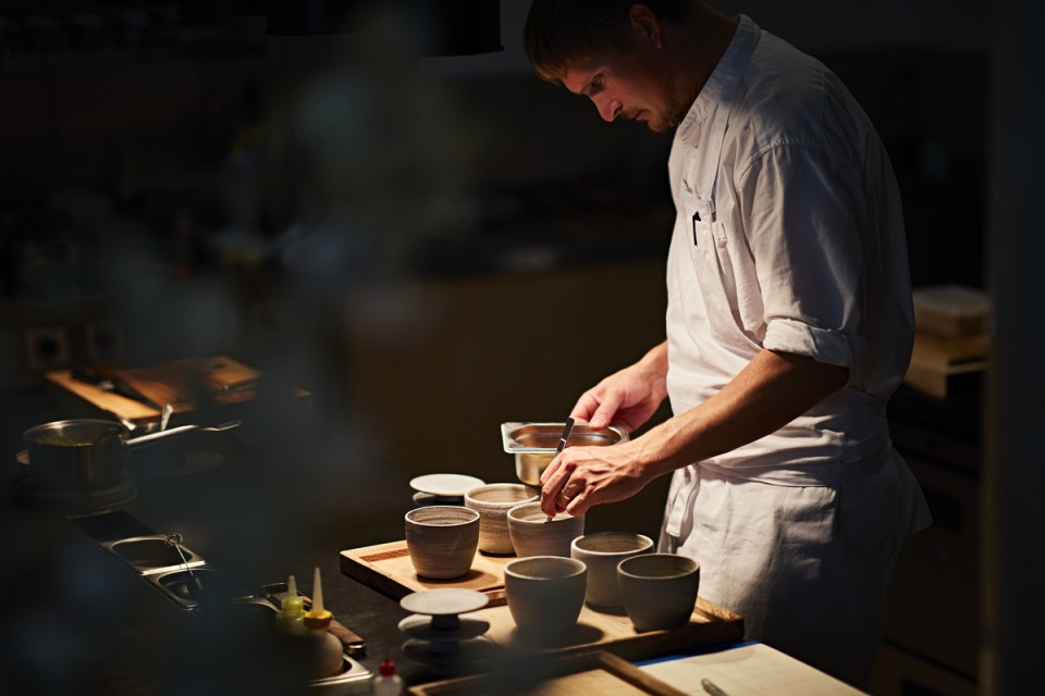 A chef in a white uniform meticulously arranging ingredients in ceramic bowls on a wooden counter in a dimly lit kitchen.