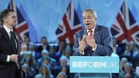 Nigel Farage speaking at a Reform UK podium, with a man in a suit and Union Jack flags in the background