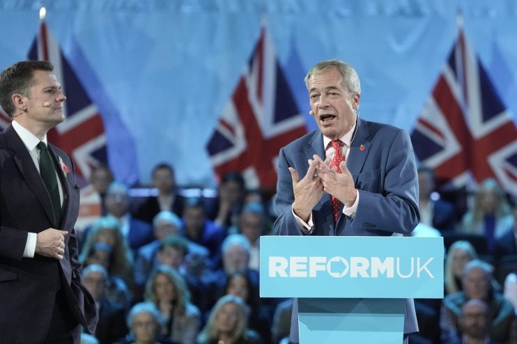 Nigel Farage speaking at a Reform UK podium, with a man in a suit and Union Jack flags in the background