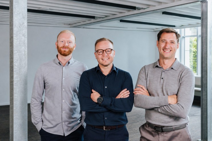Three smiling male founding team members in business casual attire standing together indoors.