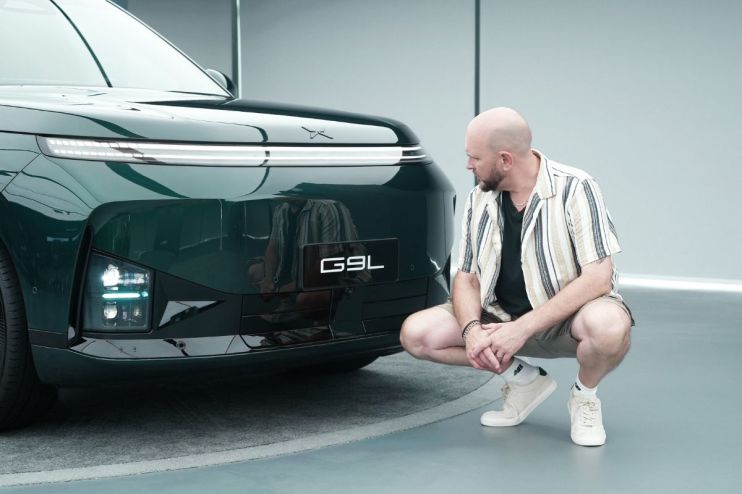 Man in striped shirt crouches next to the front of a dark green XPeng G9 electric SUV