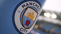 Manchester City FC logo on a light blue surface, covered in raindrops, with blurred stadium lights in background