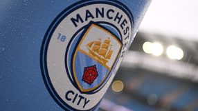 Manchester City FC logo on a light blue surface, covered in raindrops, with blurred stadium lights in background