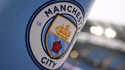 Manchester City FC logo on a light blue surface, covered in raindrops, with blurred stadium lights in background