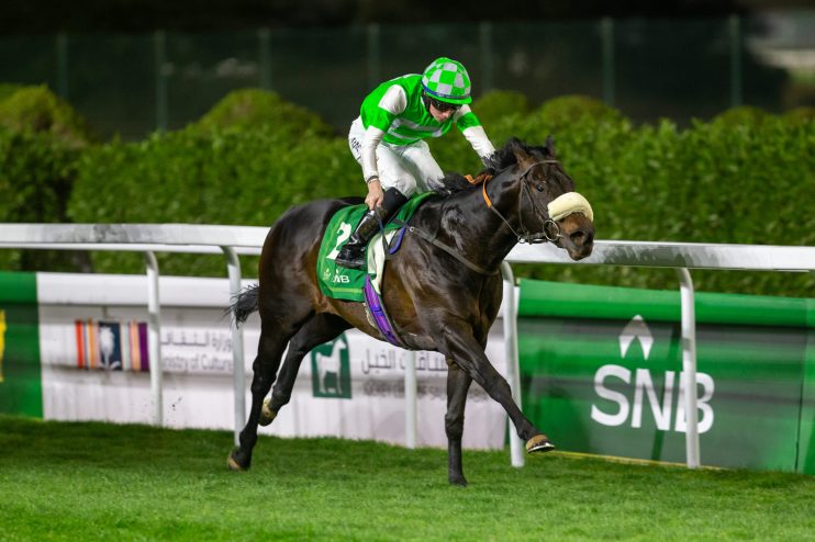 Jockey in green and white silks riding a dark brown racehorse on a green track at night