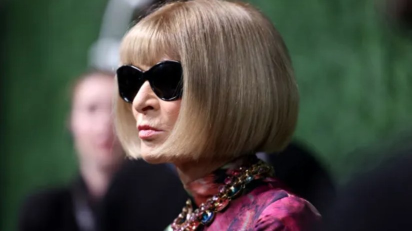 Anna Wintour in black sunglasses, blonde bob, colorful statement necklace, and a patterned top.