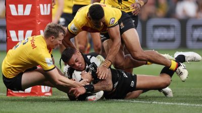 Rugby players in yellow and black jerseys tackle a player in a black and white jersey, holding a ball.