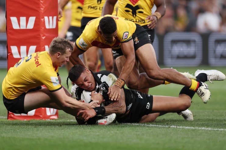Rugby players in yellow and black jerseys tackle a player in a black and white jersey, holding a ball.